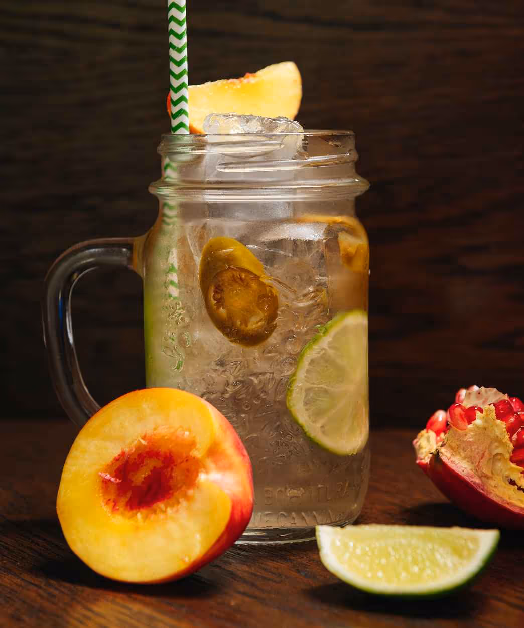 Mason jar filled with ice, lime slices, and jalapeño slices, garnished with a peach slice and green zig-zag straw, surrounded by peach, lime wedge, and pomegranate on wooden surface.